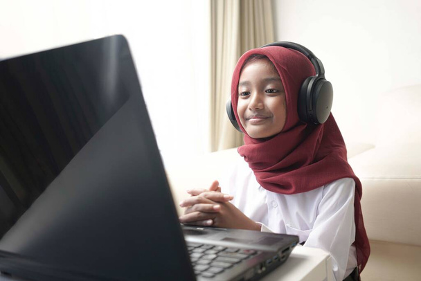 a cute little girl wearing red hijab doing something on her laptop with headphone - Foto, immagini