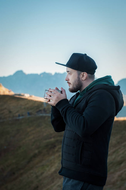 Бородатый путешественник пьет чай или кофе из металлической чашки, отдыхая на скале на красивом ландшафтном фоне - Фото, изображение