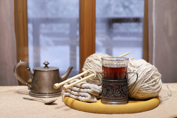 Ein Glas Tee, Wollfäden und Strickwaren auf dem Tisch vor dem Fenster vor dem Hintergrund der Winterlandschaft.  - Foto, Bild