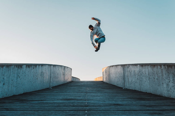 Young caucasian man in jeans and hoodie jumping on concrete bridge. Mid air parkour pose in city environment and clear sky - Фото, изображение