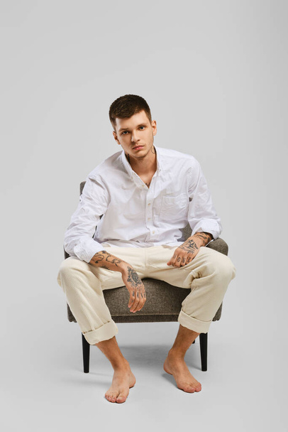 Full length studio portrait of young barefoot man in white shirt and ivory trousers sitting in armchair - Zdjęcie, obraz