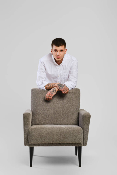 Full length studio portrait of young barefoot man in white shirt and ivory trousers leaning to armchair - Foto, imagen