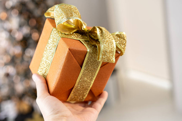 Man holding orange Christmas gift box with gold ribbon in hand with Christmas tree on background Surprise present box winter holiday - Photo, image