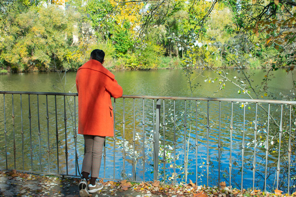 kobieta w pomarańczowym płaszczu pochylona nad poręczą rzeki - Zdjęcie, obraz