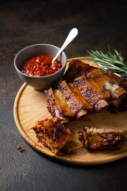 Close up of grilled ribs and bbq sauce on wooden plate on dark surface - Fotografie, Obrázek