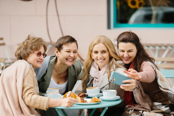 Trois caucasiennes jolies amies âgées assises avec un café à table sur la terrasse du café et souriant à la caméra du smartphone tout en prenant des photos selfie. Vieilles dames posant pour téléphoner. Pensionnés au repos. - Photo, image
