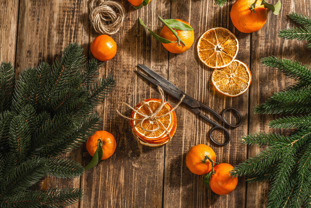 Suche pomarańcze kroją przekąski. Suszone owoce cytrusowe do dekoracji świątecznych. Mandarynki z liśćmi, gałązkami jodły, nożyczkami i nitką. Drewniane tło, koncepcja rustykalna, widok z góry - Zdjęcie, obraz