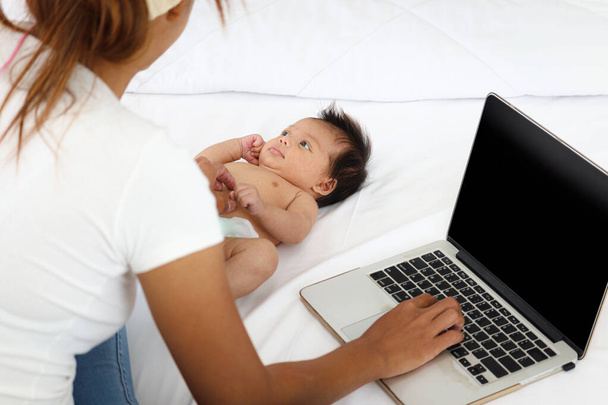 Imádnivaló kis édes újszülött kislány feküdt a fehér ágyban, közel az anyja segítségével notebook laptop számítógép, aranyos csecsemő vele elfoglalt anya, akinek otthon kell dolgoznia. - Fotó, kép