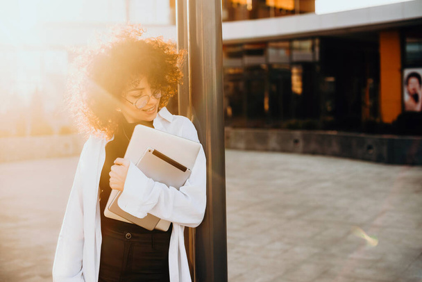 Portrait of a curly woman leaning on the metal bar and holding a laptop while waiting for someone. Sunshine. Businesswoman portrait. Curly hair. - Photo, image