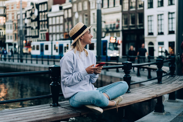 Full body of female tourist with legs crossed text messaging on mobile phone and enjoying vacation in Amsterdam while looking away - Photo, image