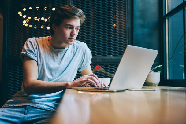 Concentrated youthful male employee wearing casual clothes sitting at wooden table with laptop and smartphone while doing freelance project in cozy restaurant - Фото, зображення