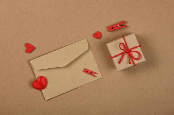 Закройте подарочную коробку от Валентина с красным бантом из бечевки и конвертом для открыток с деревянными прищепками и сердечками на коричневом бумажном фоне, плоский уголок, возвышенный вид сверху, прямо над - Фото, изображение