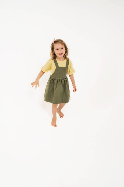A girl in a green sundress. A beautiful girl rejoices and jumps. A child with blonde hair on a light background. Real childrens emotions in a child. - Foto, immagini
