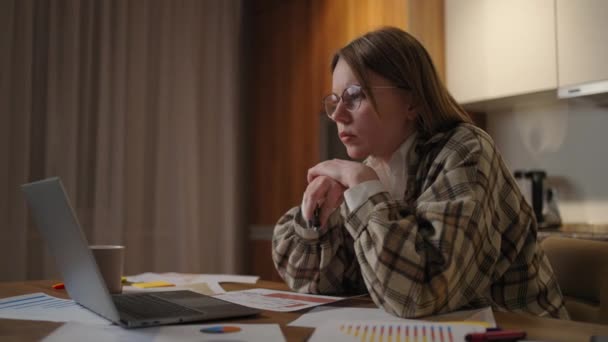 A pensive woman in glasses looks at the laptop screen and concentrates and comes up with a development strategy. Schedules and plans on paper are on the table - Footage, Video