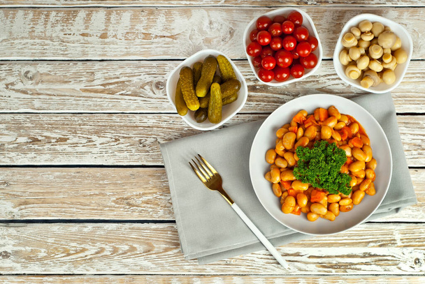 Beans with carrots in a white bowl. Beans with pickled tomatoes, mushrooms and cucumbers on a white background. Food on a shabby table. Bowl on an old wooden board. - Photo, Image