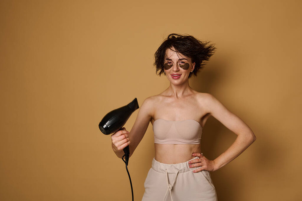Waist-length portrait on beige background of brunette a pretty with collagen spots under her eyes, directing a stream of warm air from a hairdryer to her wet hair and drying it, smiling at the camera - Photo, Image