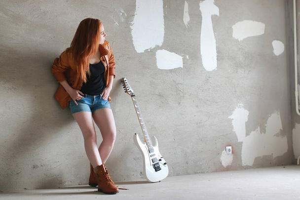 Young red-haired girl with electric guitar. Rock musician girl in a leather jacket. She is a pretty singer and a performer of rock music - Photo, image
