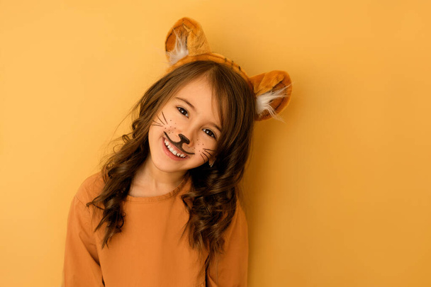 Nettes lustiges lachendes Mädchen, verkleidet als Tiger mit gemaltem Gesicht und Hut mit Ohren, isoliert auf Studiohintergrund. Ein Kind im Bild eines Tieres. Faschingskostüm. - Foto, Bild