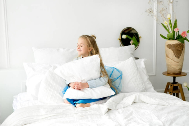 Een schattig blond meisje met lang haar zit in een bed met sneeuwwit beddengoed, knuffelt een kussen en lacht vrolijk. Comfortabel zacht beddengoed. Baby in de slaapkamer - Foto, afbeelding