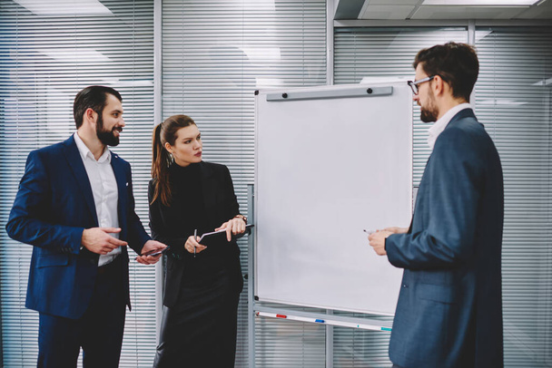 Group of business colleagues in formal clothes standing near empty whiteboard and discussing ideas of company strategy in modern office - Foto, Imagen