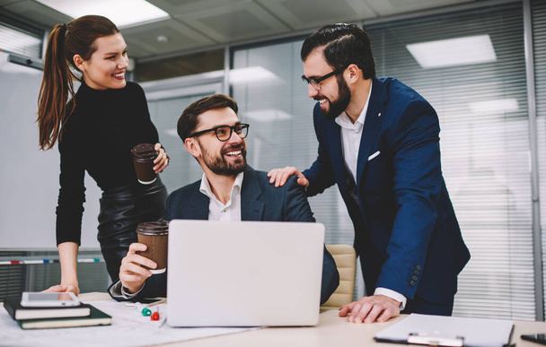 Happy coworkers in office suits looking at each other supporting man sitting at table by holding shoulder and talking in modern workspace - Foto, immagini
