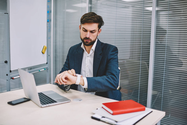 Bearded concentrated male manager in formal suit attentively looking at wristwatch while working on project on laptop in modern office - Foto, Imagem