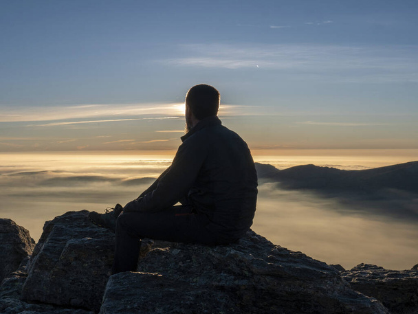 Una vista de silueta de un macho caucásico sentado en la cima de una montaña mirando hacia el atardecer - Foto, Imagen