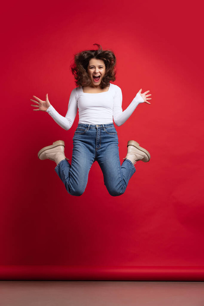 Full-length portrait of excited young beautiful girl in casual clothes jumping isolated on red color background - 写真・画像
