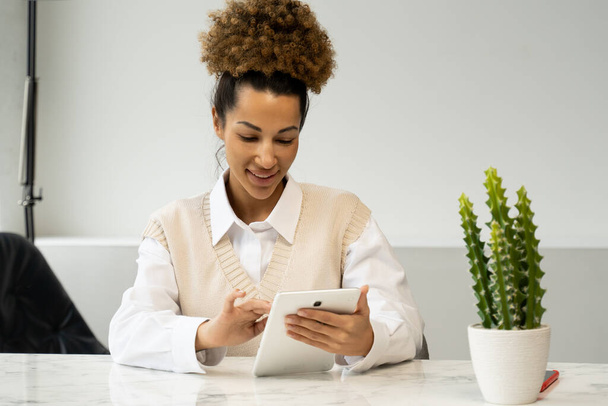 Portrait of a cheerful stylish black woman in casual clothes sitting at a desk using a tablet in the office. - Photo, image