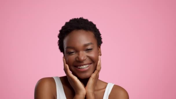 Femme noire avec des bretelles souriant toucher le visage sur fond rose - Séquence, vidéo