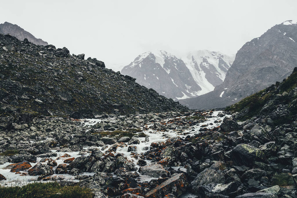 Ατμοσφαιρικό τοπίο με βουνίσιο ρυάκι μεταξύ των Μοραΐνων σε βροχερές καιρικές συνθήκες. Bleak τοπίο με γαλακτώδες ποτάμι από χιονισμένα βουνά. Πέτρες με βρύα και λειχήνες στο ρυάκι του νερού. Ορεινός ποταμός ανάμεσα σε βράχια. - Φωτογραφία, εικόνα