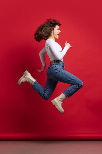 Dynamic portrait of excited young beautiful girl in casual clothes jumping isolated on red color background - Fotoğraf, Görsel