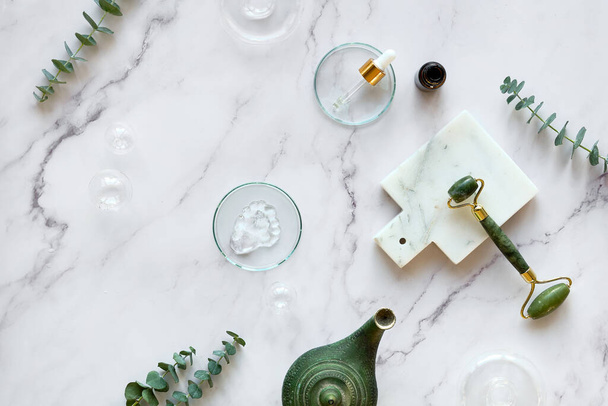 Handmade moisturizer and green jade face roller. Cotton towel. Exotic eucalyptus leaves and water drops on off white marble background with stone cutting board and oil lamp. - Foto, Imagem