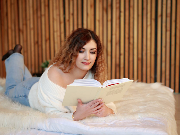 Beautiful young woman reading a book indoors, lying on the couch - Фото, изображение