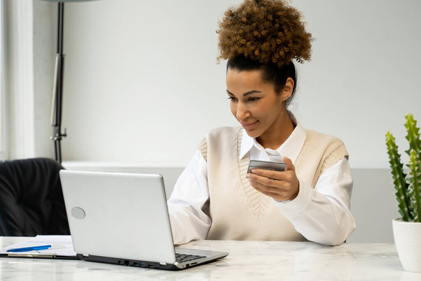 African-American woman holding a credit card, sitting at a table with a laptop, shopping online, making Internet payments - 写真・画像