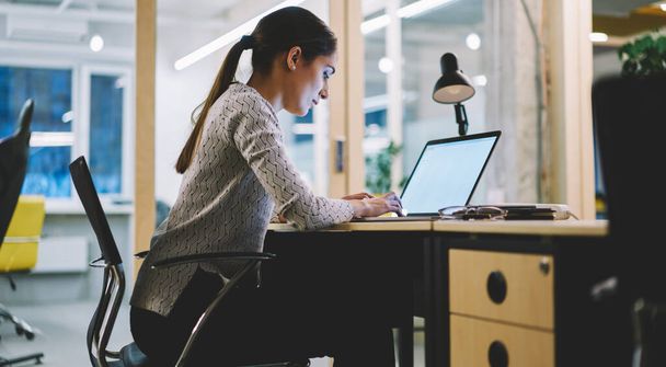 Side view of focused woman in casual clothes with dark hair sitting at desk and typing on netbook in modern workspace - 写真・画像