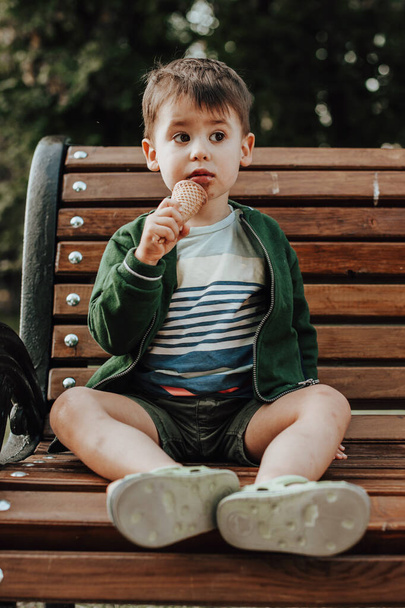 公園のベンチで手にアイスクリームを持っている白人の少年の完全な長さ。アイスクリームを食べる。白人の少年. - 写真・画像