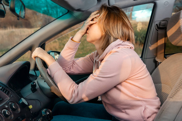 Μια νεαρή ξανθιά λυπημένη γυναίκα κάθεται πίσω από το τιμόνι ενός αυτοκινήτου και κρατά το κεφάλι της σε απόγνωση, καλύπτοντας τα μάτια της με το χέρι της. Πλευρική άποψη. Άγχος κατά την οδήγηση ενός δεξιού αυτοκινήτου. - Φωτογραφία, εικόνα