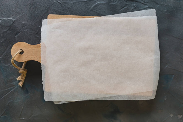 Дерев'яна обробна дошка та білий пергаментний папір на темно-сірому фоні. Макет рецептів, списку покупок або їжі
 - Фото, зображення