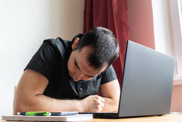 pääaihe on epätarkka, nuori mies istuu vieressä pöydällä, kotitoimisto verkossa kannettava tietokone työ liike etäisyys freelancer kyllästynyt väsynyt onneton - Valokuva, kuva