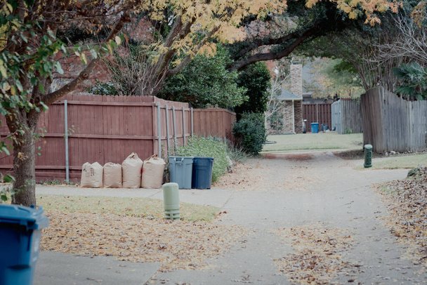 Tönendes Foto einer ruhigen Seitengasse mit einer schönen Decke aus abgefallenen Blättern in einem Vororthaus in der Nähe von Dallas, Texas, Amerika. Reihe von Papier-Rasensäcken und Mülltonnen entlang des Holzzaunes - Foto, Bild