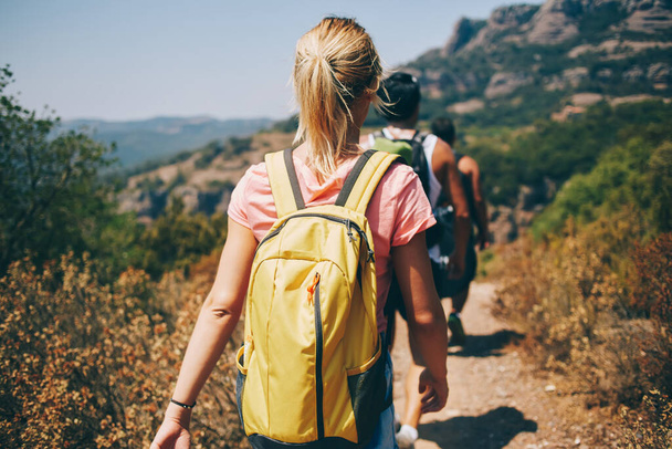 Back view of young female backpacker hiking with group of tourists on sandy road in mountain terrain in sunny summer day - Foto, imagen