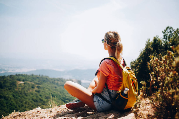 Full body back view of faceless young female tourist admiring picturesque view while wearing summer outfit and sunglasses while sitting on hilltop with backpack - Фото, изображение