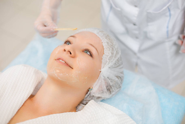 Hands of cosmetology specialist applying facial mask using stick, making skin hydrated and face glowing. Attractive woman relaxing smiling and enjoying spa procedures - Photo, image