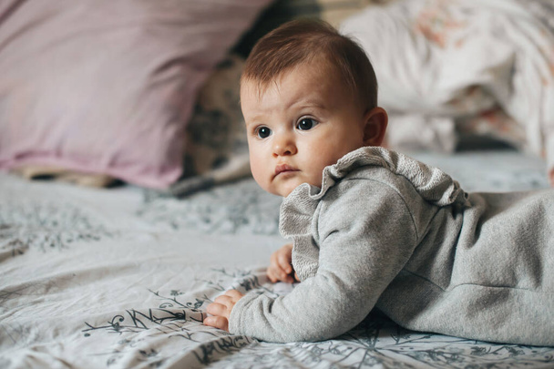 Die Seitenansicht eines kleinen Mädchens liegt auf dem Bauch auf dem Bett und blickt in die Kamera. Neugeborenes. Schöne Mädchen. Gesunder Lebensstil. - Foto, Bild