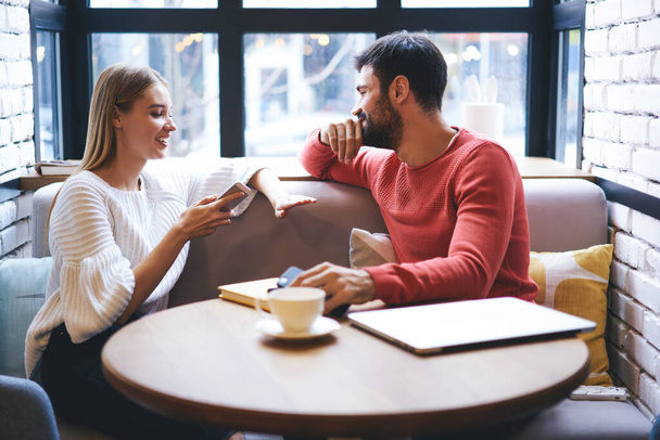 Young attractive woman telling interesting story written on smartphone to boyfriend while relaxing together during coffee break in cozy comfortable cafeteria - Photo, Image
