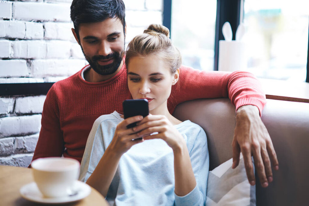 Calm couple in casual clothes sitting at table with cup of drink and using smartphone while chilling together in cafe - Photo, Image