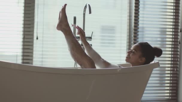 Depilación en casa. Joven mujer afroamericana positiva afeitándose las piernas con navaja de afeitar, relajándose en el baño en casa - Metraje, vídeo