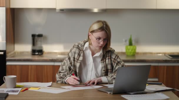 Una mujer con gafas trabaja remotamente desde casa sentada en una mesa con un portátil y un rotulador marca los datos en el gráfico. Estudiante de educación a distancia en casa para realizar una tarea en economía - Imágenes, Vídeo