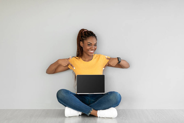 Pretty young black woman sitting cross legged on floor, pointing at empty laptop with mockup for website design - Photo, Image